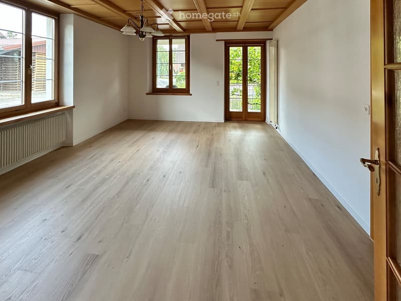 Empty room with light wooden flooring, white walls, large windows, and a wooden door leading outside.