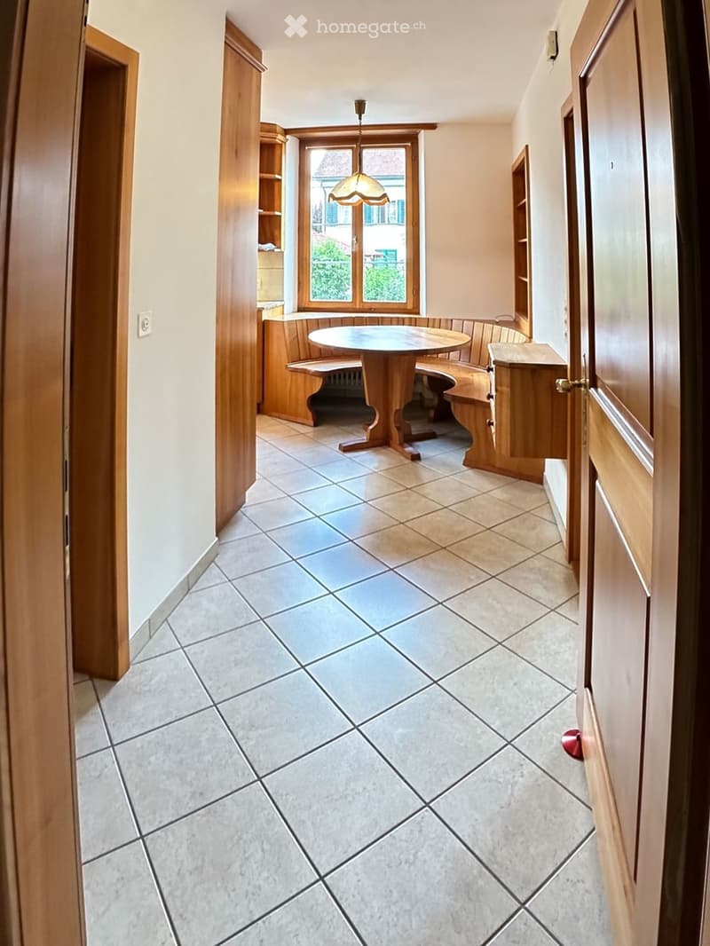 A hallway with tiled flooring, wooden doors, and a small round table with benches in the background, illuminated by natural light from a window.