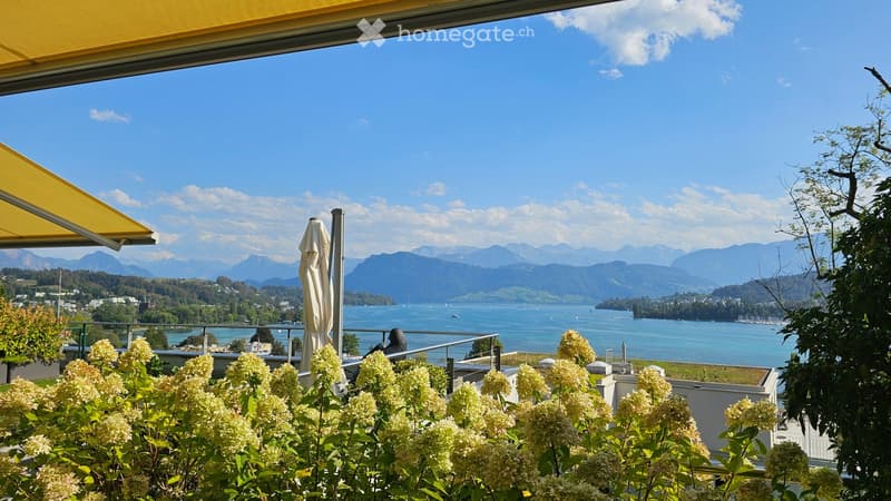 Balcony with a view of a lake and mountains, featuring a yellow awning, a closed umbrella, and flowering shrubs in the foreground.