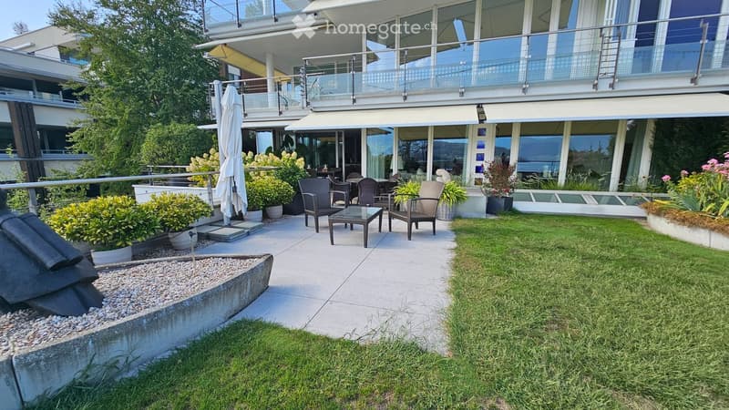 Well-maintained garden area with a seating set, including chairs and a table, surrounded by greenery and a modern building with large windows and a balcony.