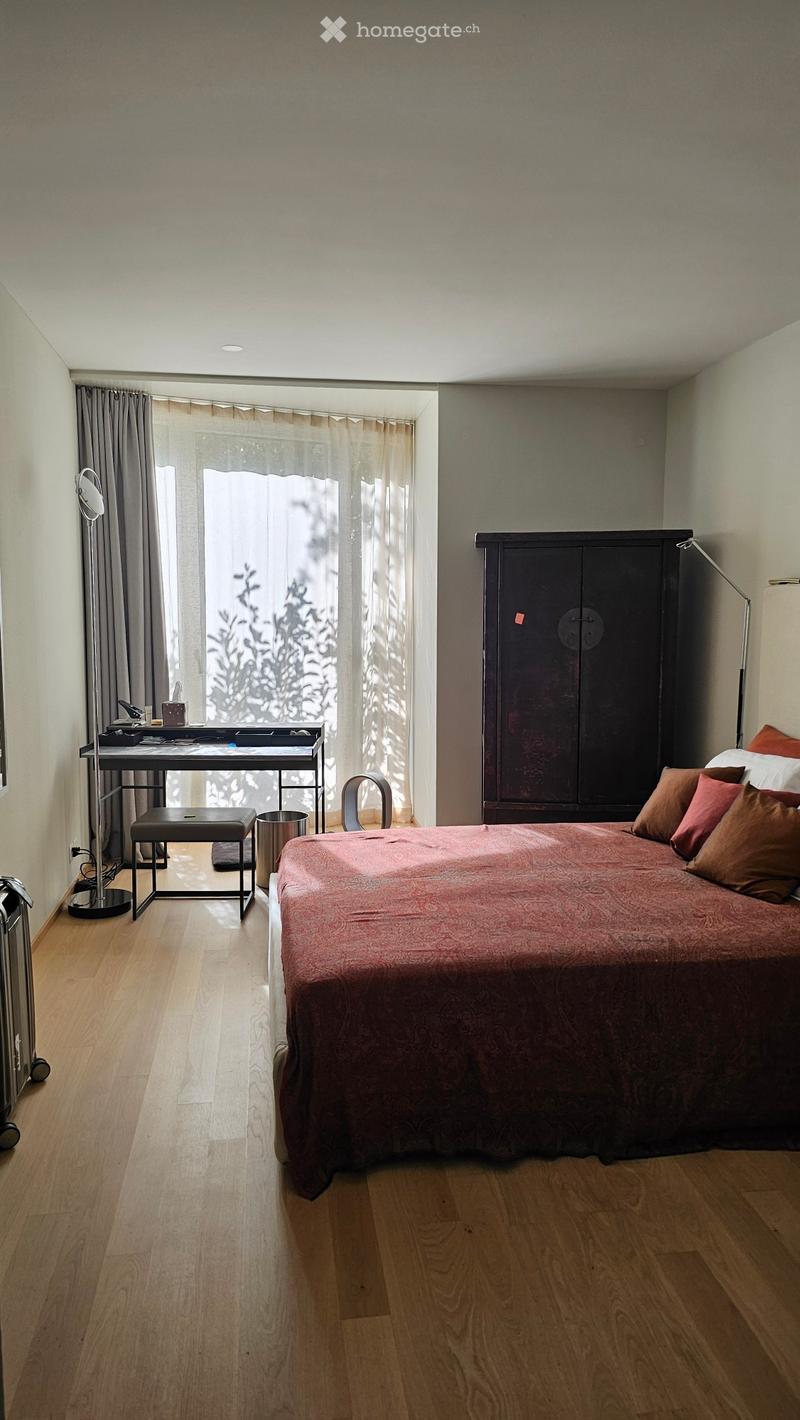 Modern bedroom with wooden flooring, large window with sheer curtains, bed with patterned red duvet and pillows, dark wardrobe, desk with chair, and floor lamp.
