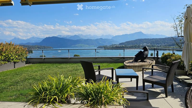 Balcony with a scenic view of a lake and mountains, featuring a small seating area with a table and chairs, surrounded by greenery and a decorative bird sculpture.