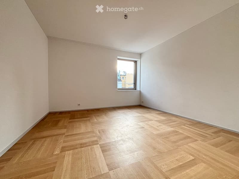 Empty room with light wooden flooring and white walls, featuring a single window with a view of a building outside.