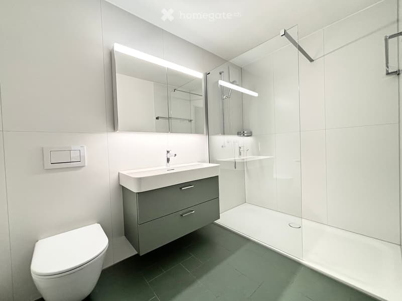 Modern bathroom with white tiles, a toilet, a sink with a dark grey cabinet, a large mirror, and a glass-enclosed shower area.