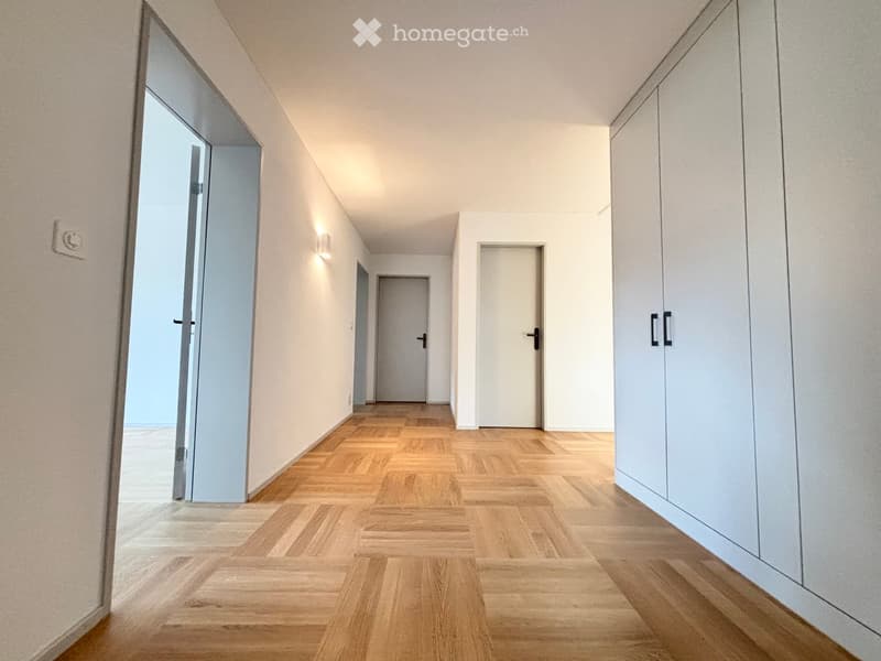 A modern hallway with light wooden herringbone flooring, white walls, and several closed doors. The hallway features built-in white cabinets on one side and recessed lighting.