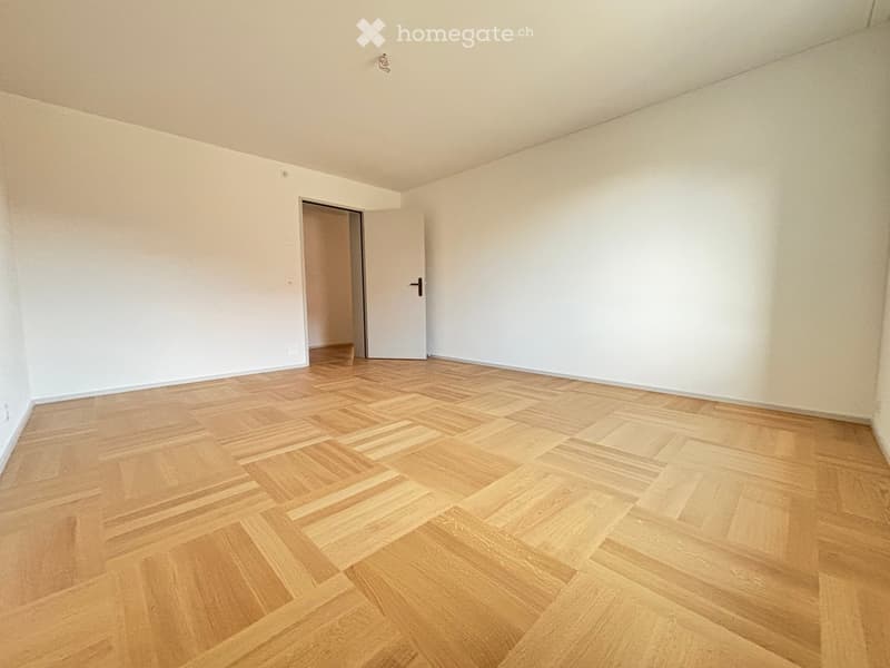 Empty room with light wooden parquet flooring and white walls, featuring a single door leading to another space.