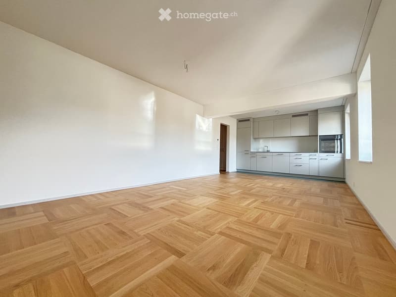 Empty room with light-colored walls and a herringbone wooden floor, featuring a small, modern kitchen area with cabinets and appliances in the corner.