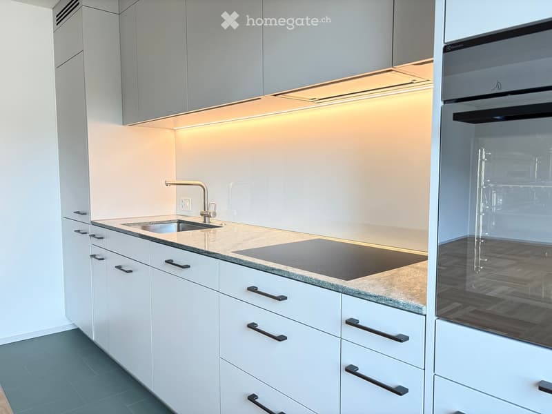 Modern kitchen with white cabinets, a sink, faucet, induction cooktop, and an oven. The countertops are light-colored with a subtle pattern, and the lighting is warm and ambient.