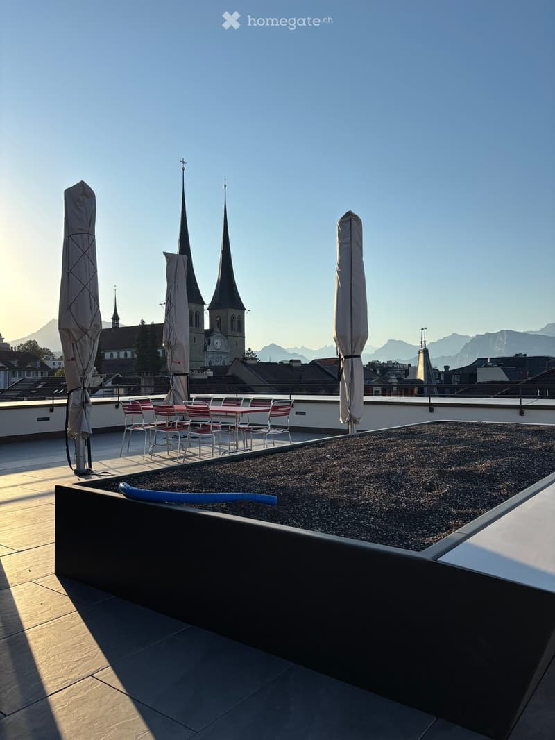 Roof terrace with seating area, closed umbrellas, and a view of a city with spires and mountains.