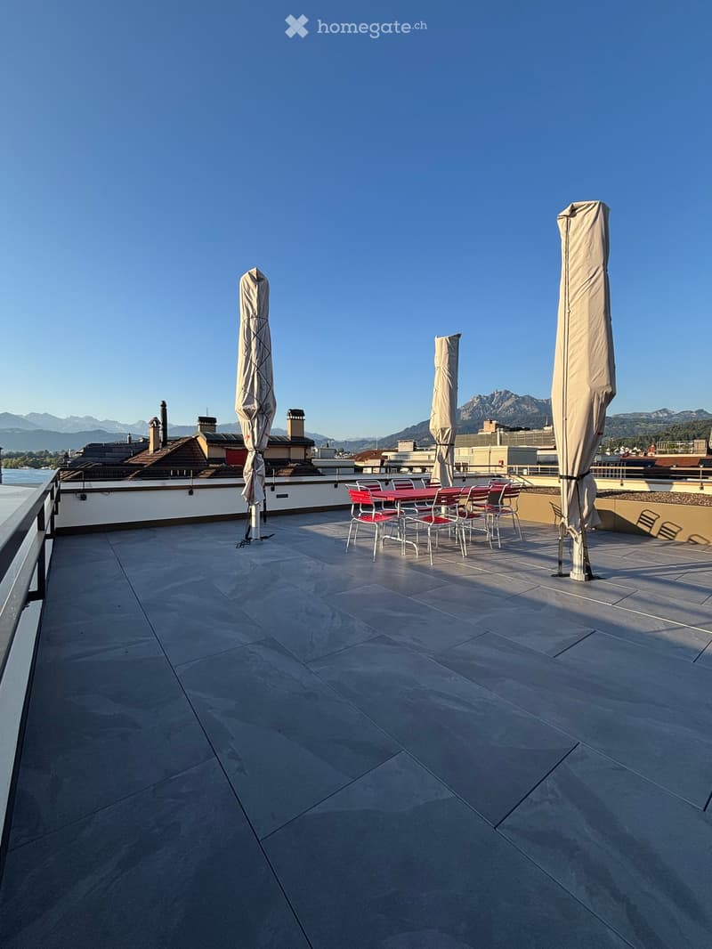 Roof terrace with stone tiles, red table, and chairs, with closed umbrellas and a scenic mountain view.