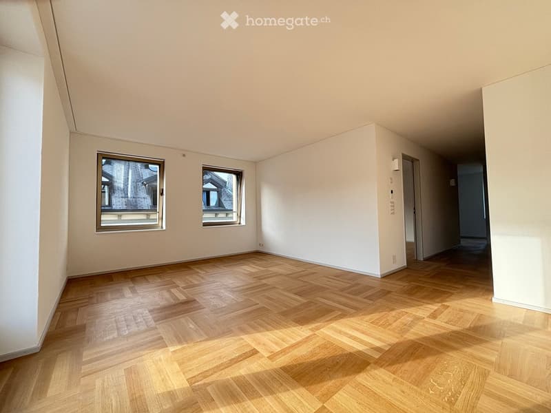 Empty room with light wooden herringbone flooring, white walls, and two windows showing a view of neighboring rooftops.