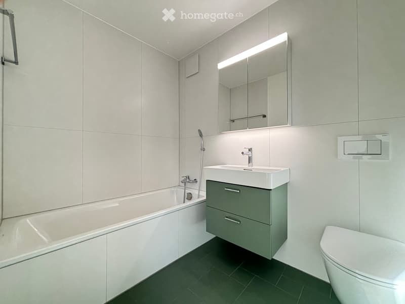 Modern bathroom with a white bathtub, a white sink with a grey cabinet, a white toilet, and a large mirror with built-in lighting.