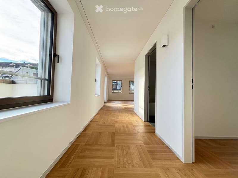 Modern hallway with light wooden flooring, white walls, large windows, and a door leading to another room.