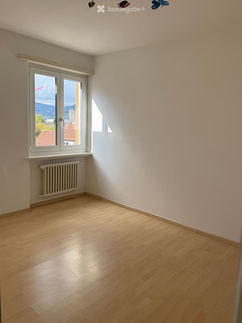Empty room with light wooden flooring, white walls, and a large window offering a view of the city and mountains.