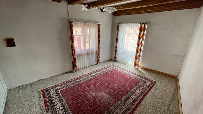 Empty room with exposed wooden beams on the ceiling, two windows with sheer curtains, and a red patterned rug on the floor.