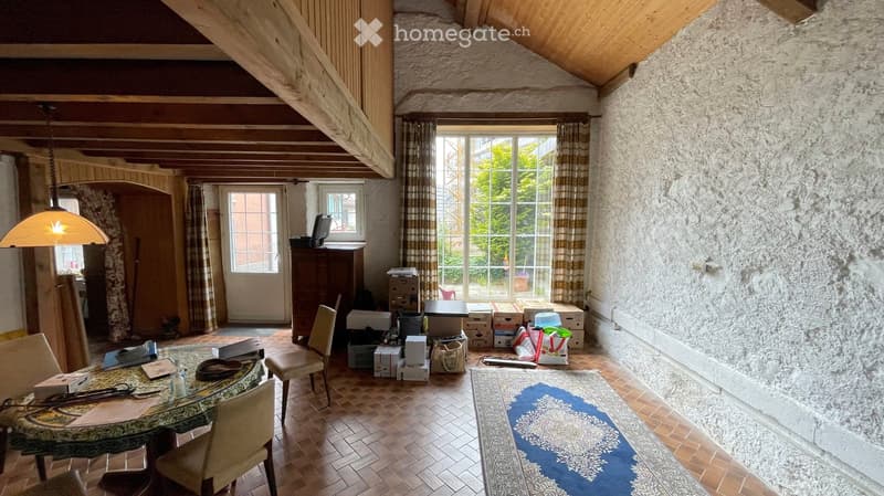A furnished room with exposed wooden beams, stone walls, and wooden flooring. Features include a round dining table with chairs, a hanging lamp, a cabinet, and a window with curtains.