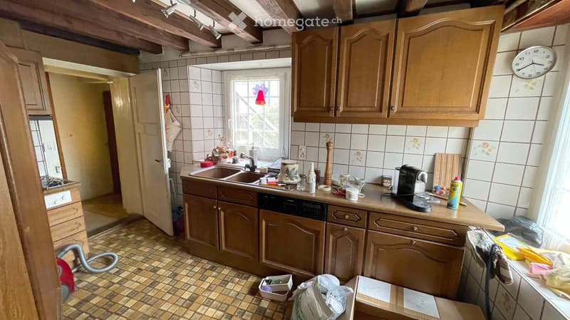 A cozy kitchen with wooden cabinets, a sink, a dishwasher, and various appliances on the counter, including a coffee maker and a bottle. The room features tiled walls and a tiled floor, with a window letting in natural light.