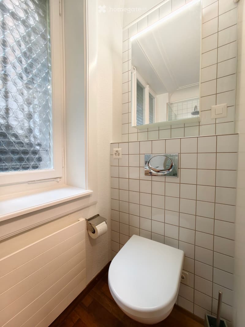 Modern bathroom with white tiles, a toilet, a mirror, a glass shelf, and a window with decorative glass.