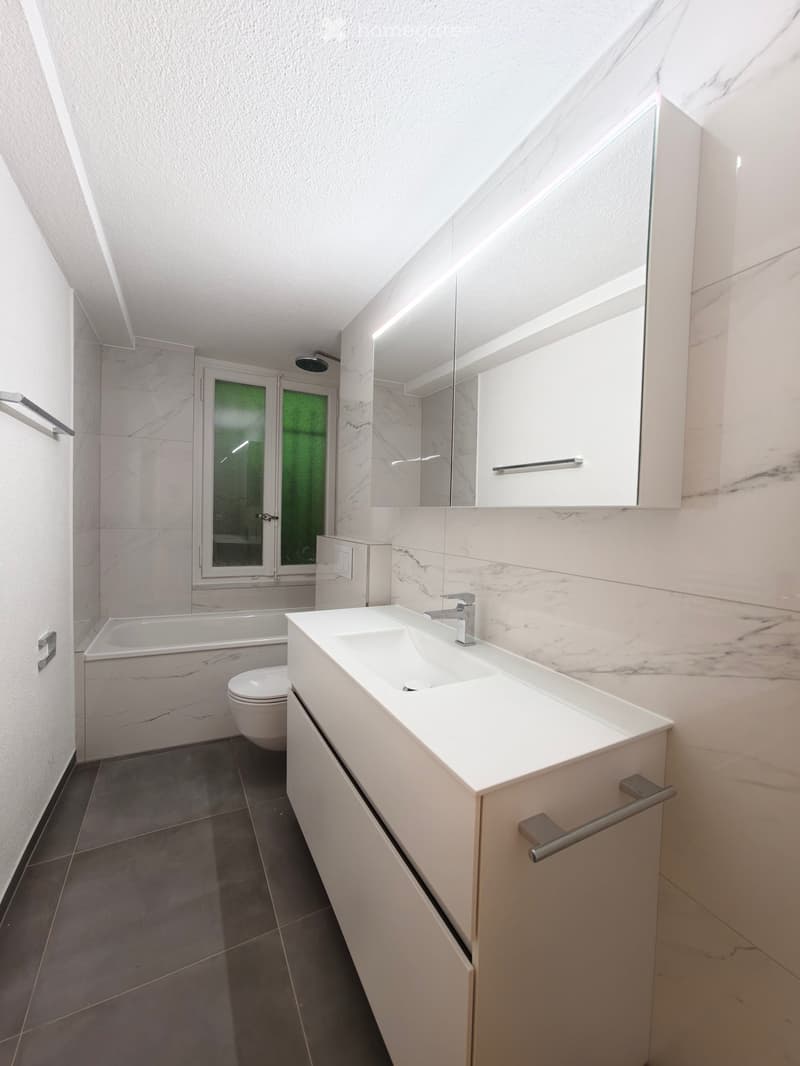 Modern bathroom with a white sink, mirror, bathtub, and toilet. The walls are tiled in a marble pattern, and the floor is dark grey tiles.