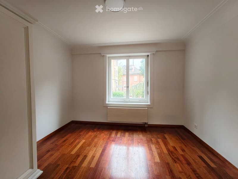 Empty room with wooden flooring, white walls, a window with white frame, and a radiator below the window.