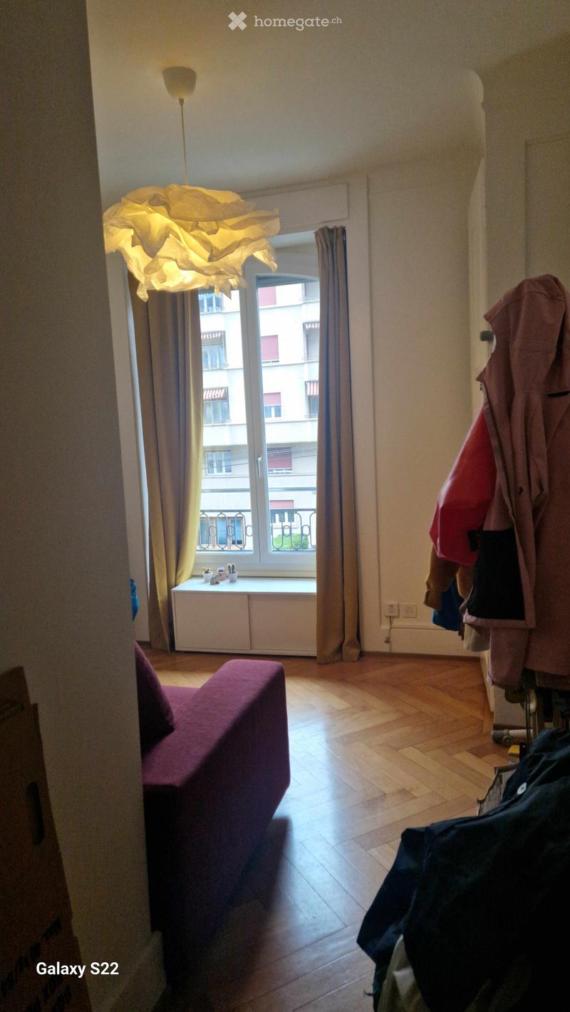 A small, well-lit living room with wooden flooring, a purple sofa, a modern lamp, a curtained window with a view of a cityscape, and a coat rack with jackets.