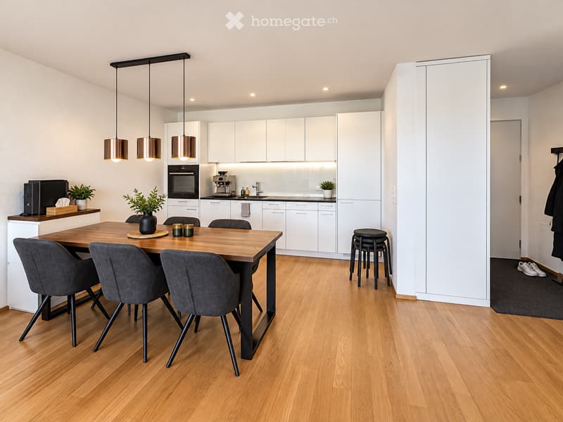 Modern kitchen and dining area with white cabinets, wooden dining table, grey chairs, pendant lights, and a small bar area with a laptop and coffee maker.