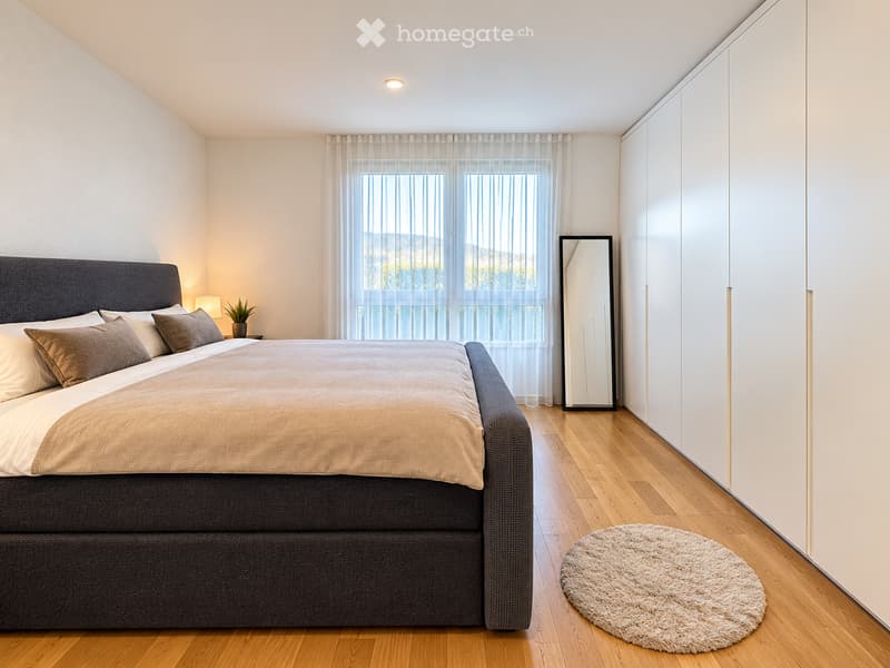 Modern bedroom with a large bed, nightstand, potted plant, mirror, and a round shaggy rug on the wooden floor.