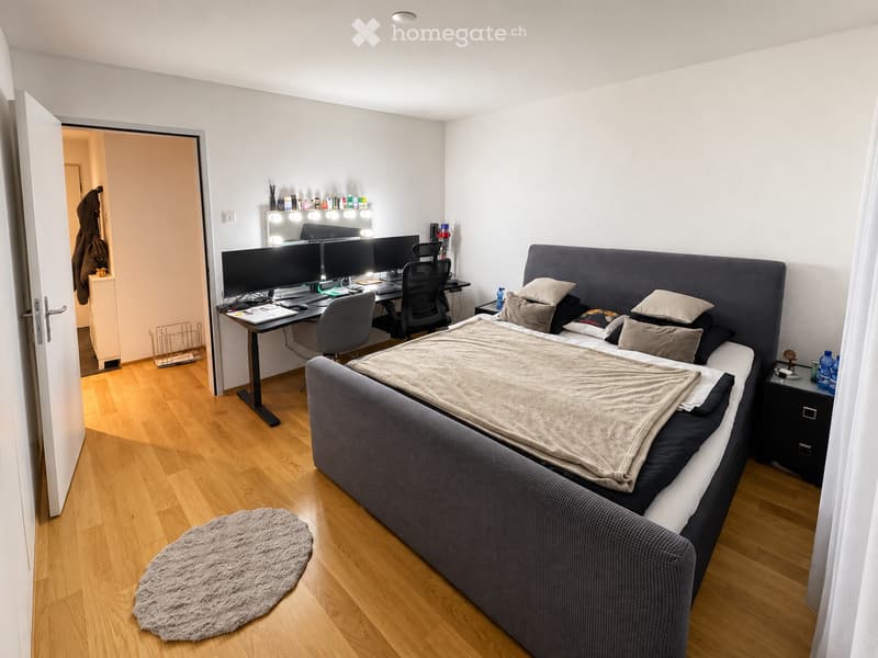Modern bedroom with a large bed, desk with multiple screens, and a nightstand. The room features wooden flooring, a round rug, and a mirror with lights above the desk.