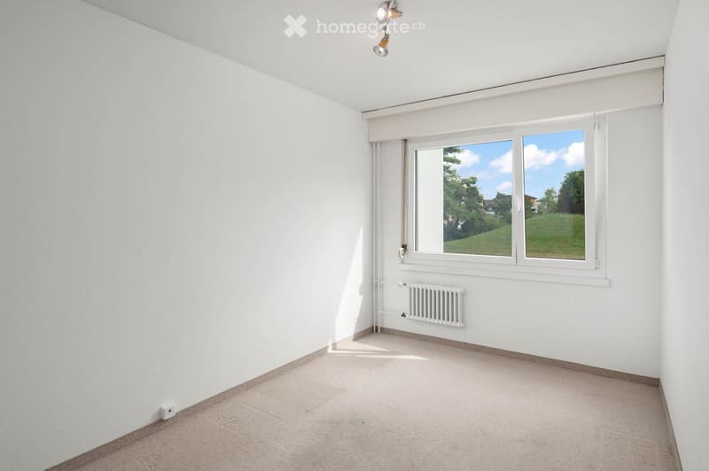 Empty room with white walls, carpeted floor, large window with blinds, radiator, and ceiling light fixture.