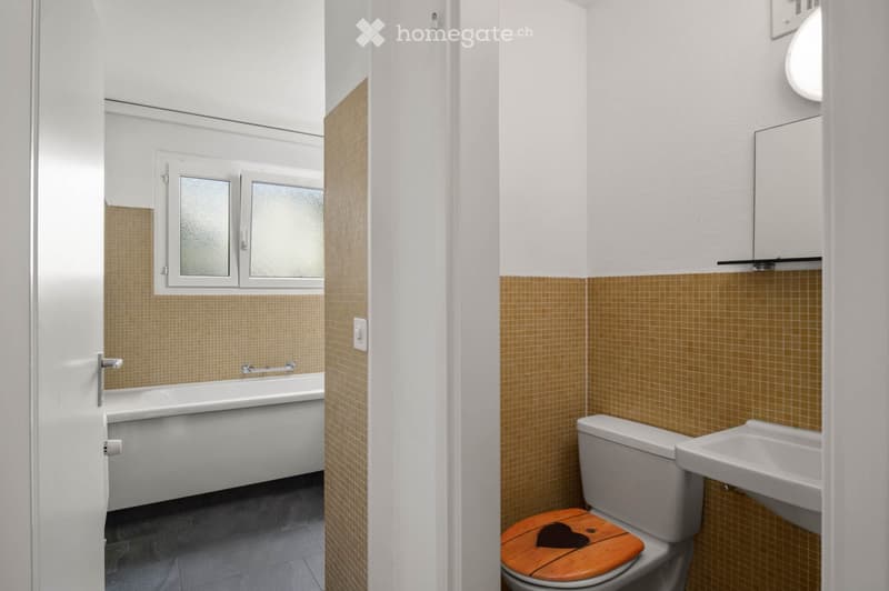 Modern bathroom with a bathtub, toilet, sink, and mirror. Features tiled walls and a wooden toilet seat with a heart design.