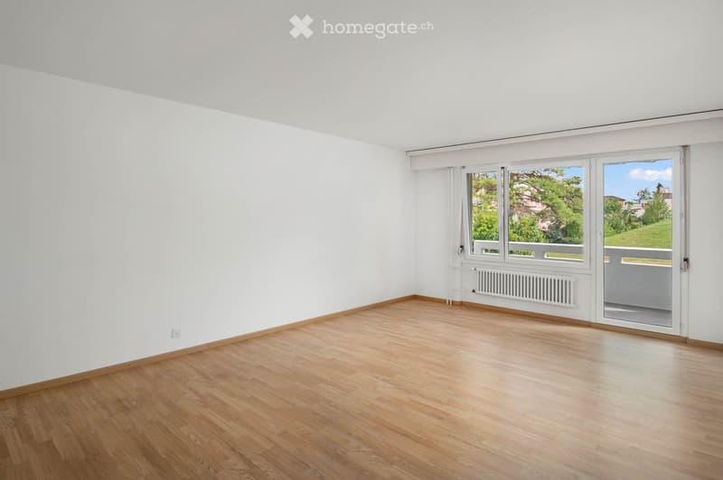 Empty room with light wooden flooring, white walls, and large windows offering a view of greenery outside. The room features a radiator and a sliding glass door leading to a balcony.