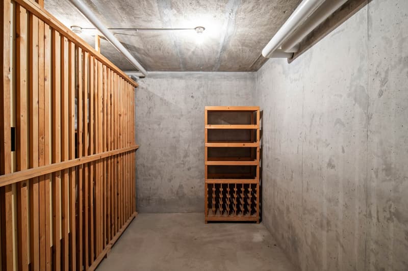 A narrow hallway with concrete walls and a wooden shelf holding wine bottles. The hallway features exposed pipes and a wooden railing on one side.