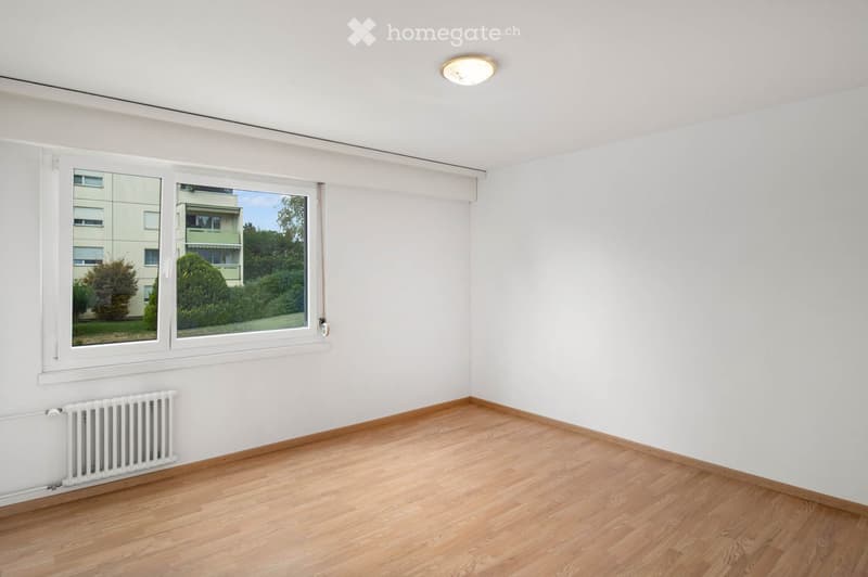 Empty room with light wooden flooring, white walls, large window with white frame, radiator, and a ceiling light fixture.