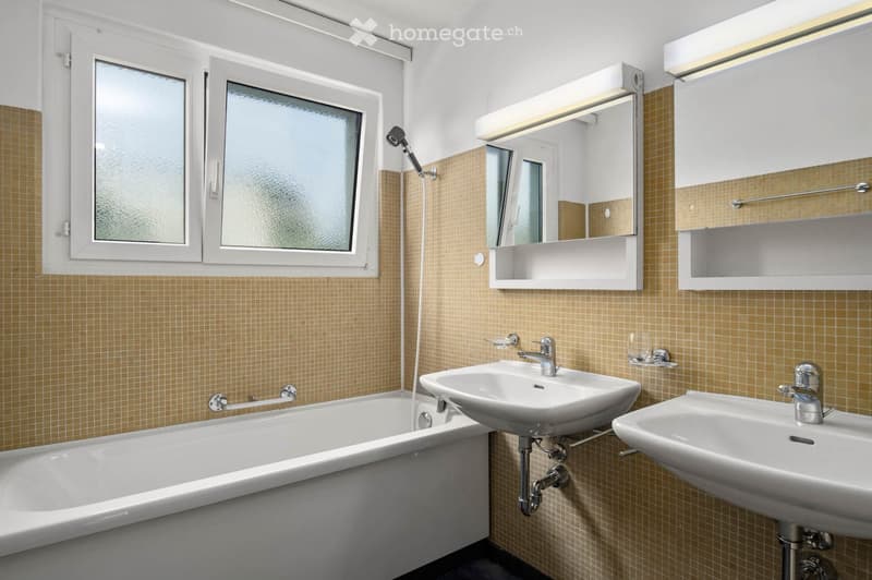 Modern bathroom with a bathtub, two sinks, a mirror, and a showerhead. The walls are tiled in a warm, beige color, and there are built-in cabinets above the sinks.