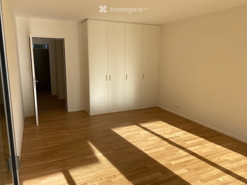 Empty room with light wooden flooring, white walls, and a large white wardrobe unit. Sunlight streams in from a window, casting shadows on the floor.