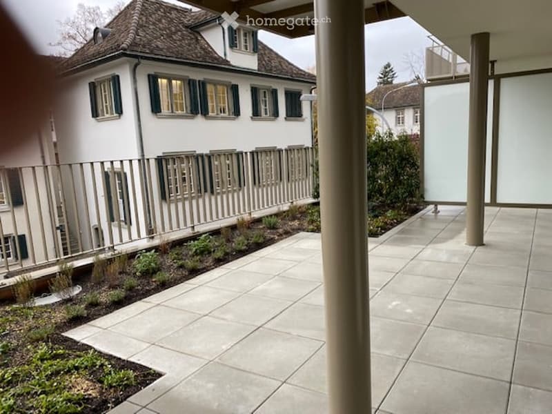 A modern patio area with a tiled floor, a metal fence, and a small garden bed with plants. The background shows a white house with green shutters and a sloped roof.