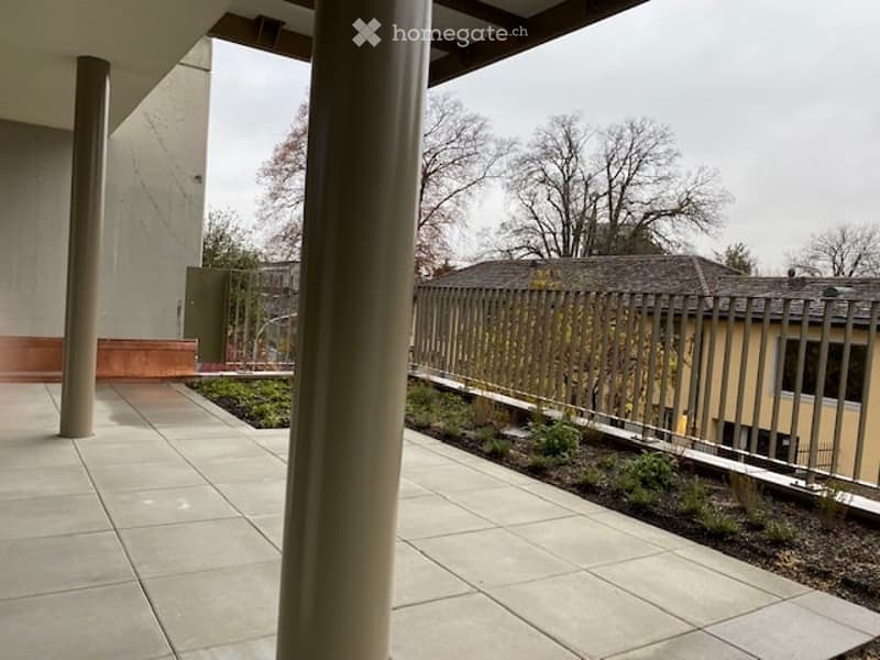 A modern outdoor patio area with tiled flooring, low metal fence, small landscaped garden beds, and surrounding buildings.