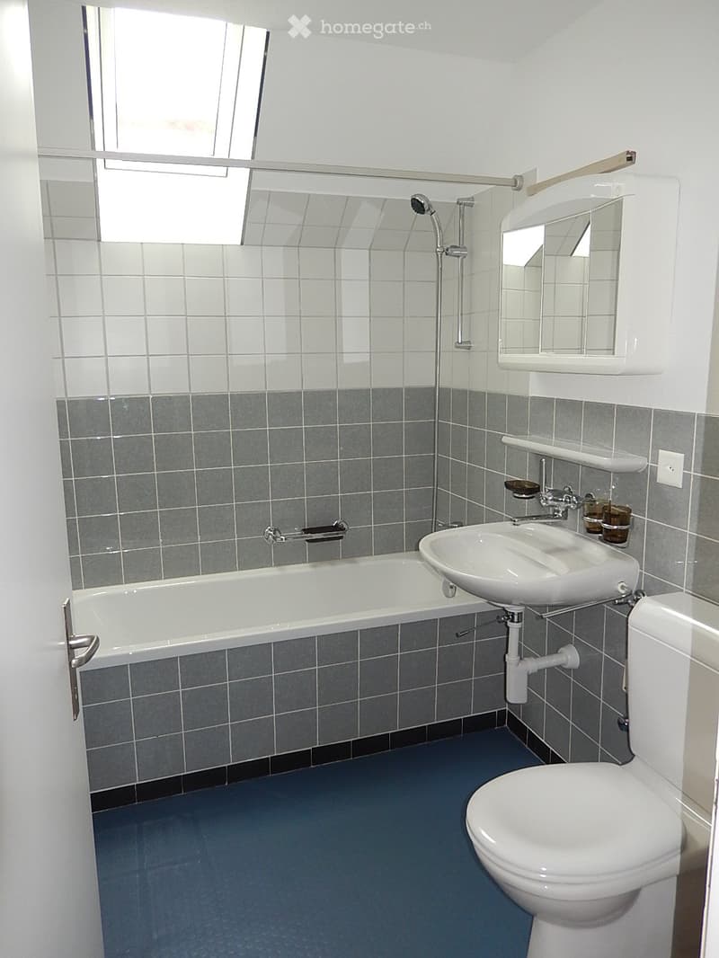 Modern bathroom with a bathtub, sink, toilet, mirror, and showerhead. Grey tiled walls and dark grey floor.