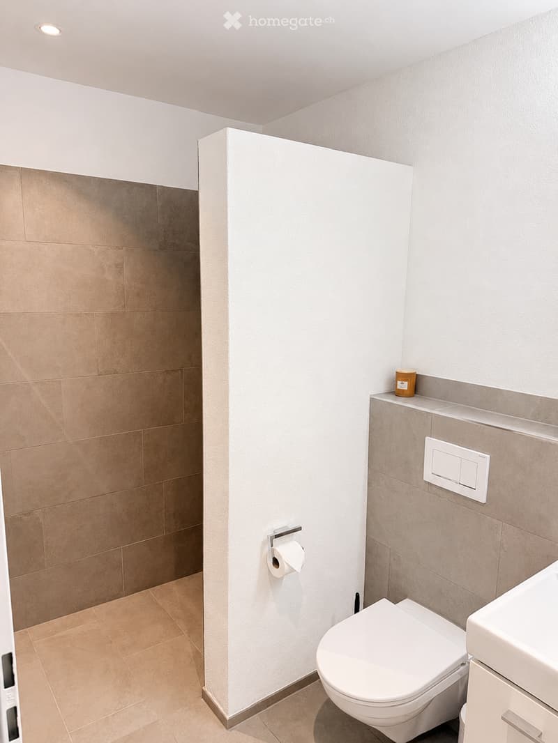 Modern bathroom with beige tiles, white toilet, sink, toilet paper holder, and a small cup on the shelf.