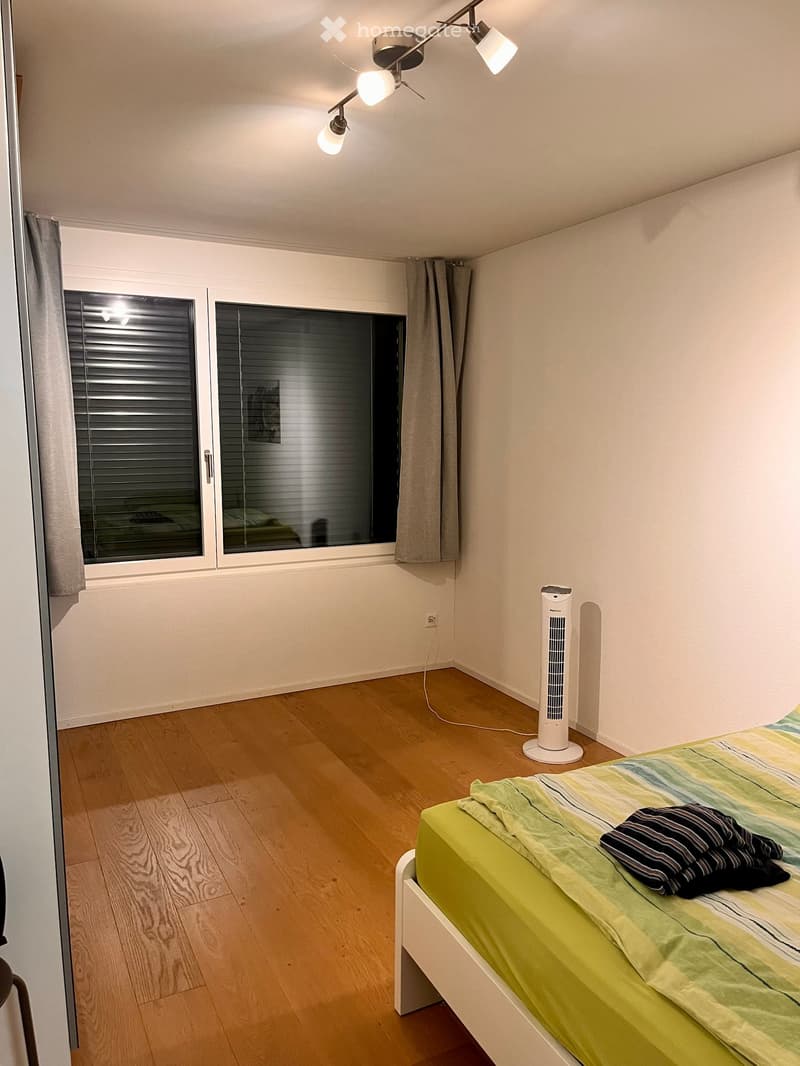 A small, tidy bedroom with a single bed, a fan, and a large window with blinds. The room features wooden flooring and white walls.