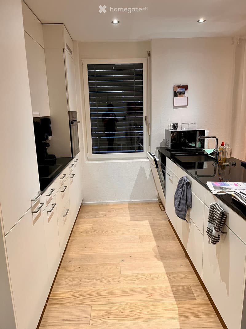 Modern kitchen with white cabinets, black countertops, and a large window with blinds. Features include a sink, microwave, oven, and various small appliances.