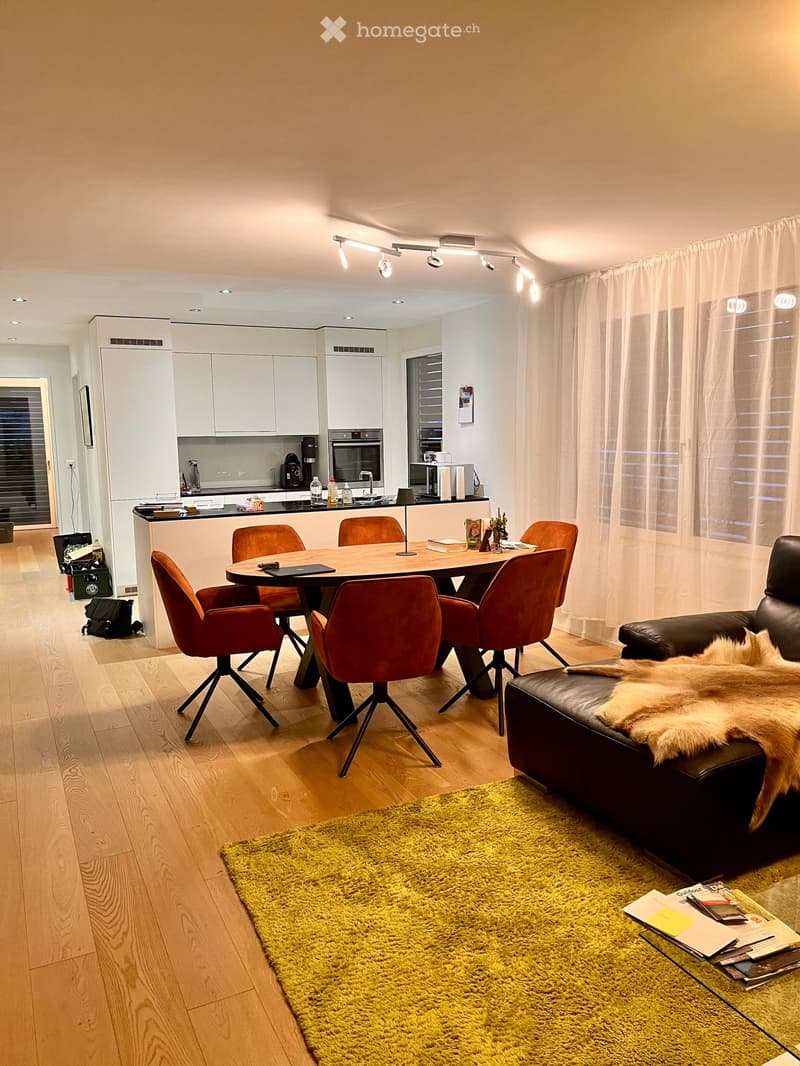 Modern open-plan living and dining area with wooden flooring, large windows with sheer curtains, a leather sofa with a fur throw, a dining table with six chairs, a kitchen area with white cabinets, an oven, and a countertop with various items.