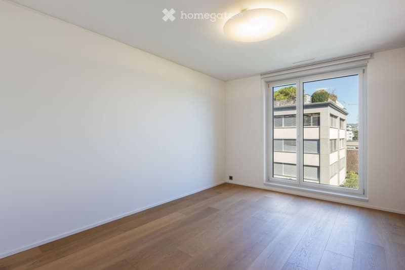 Empty room with wooden flooring, white walls, and large window with a view of neighboring buildings.