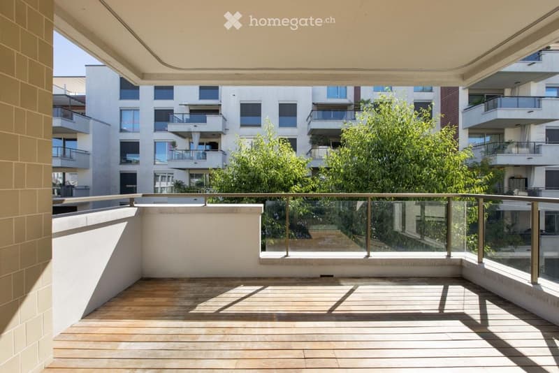 Modern balcony with wooden decking, glass railing, and view of surrounding buildings and greenery.
