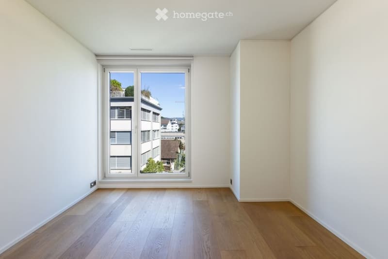 Empty room with light-colored wooden flooring and white walls, featuring a large window with a view of neighboring buildings and greenery.