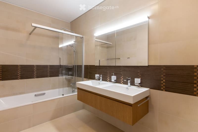 Modern bathroom with a double sink, bathtub, and glass-enclosed shower. Features include a large mirror with built-in lighting, beige tiles, and a dark brown accent border.