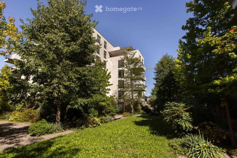 Modern multi-story residential building surrounded by lush greenery and trees, with balconies visible on the upper floors.
