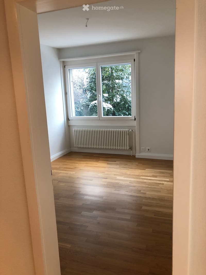 Empty room with wooden flooring, white walls, a radiator, and a large window with white frame showing greenery outside.