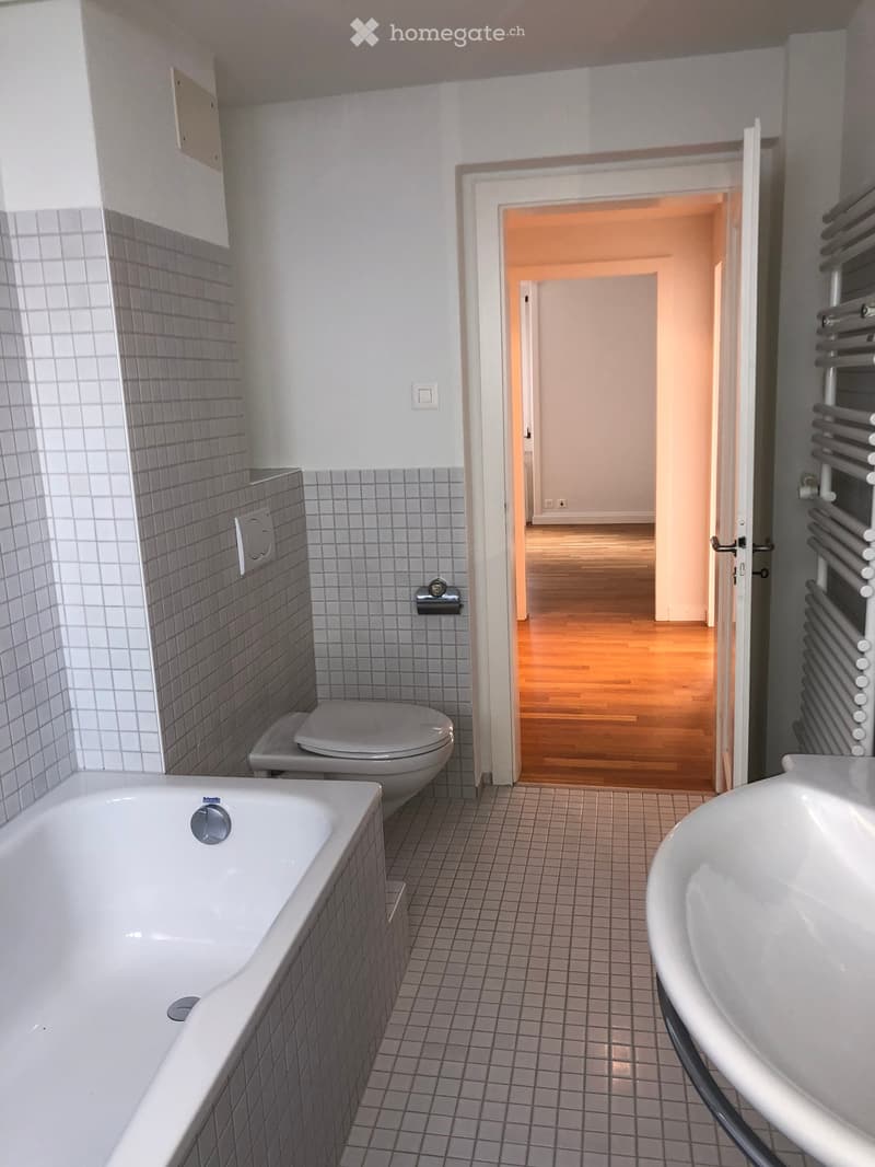 Modern bathroom with a bathtub, toilet, sink, and radiator. The walls are tiled, and there is an open doorway leading to another room with wooden flooring.