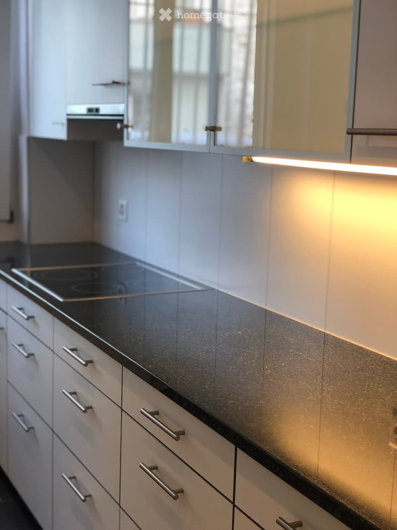 Modern kitchen with white cabinets, black countertop, and built-in induction cooktop. Features sleek stainless steel handles and ambient lighting.